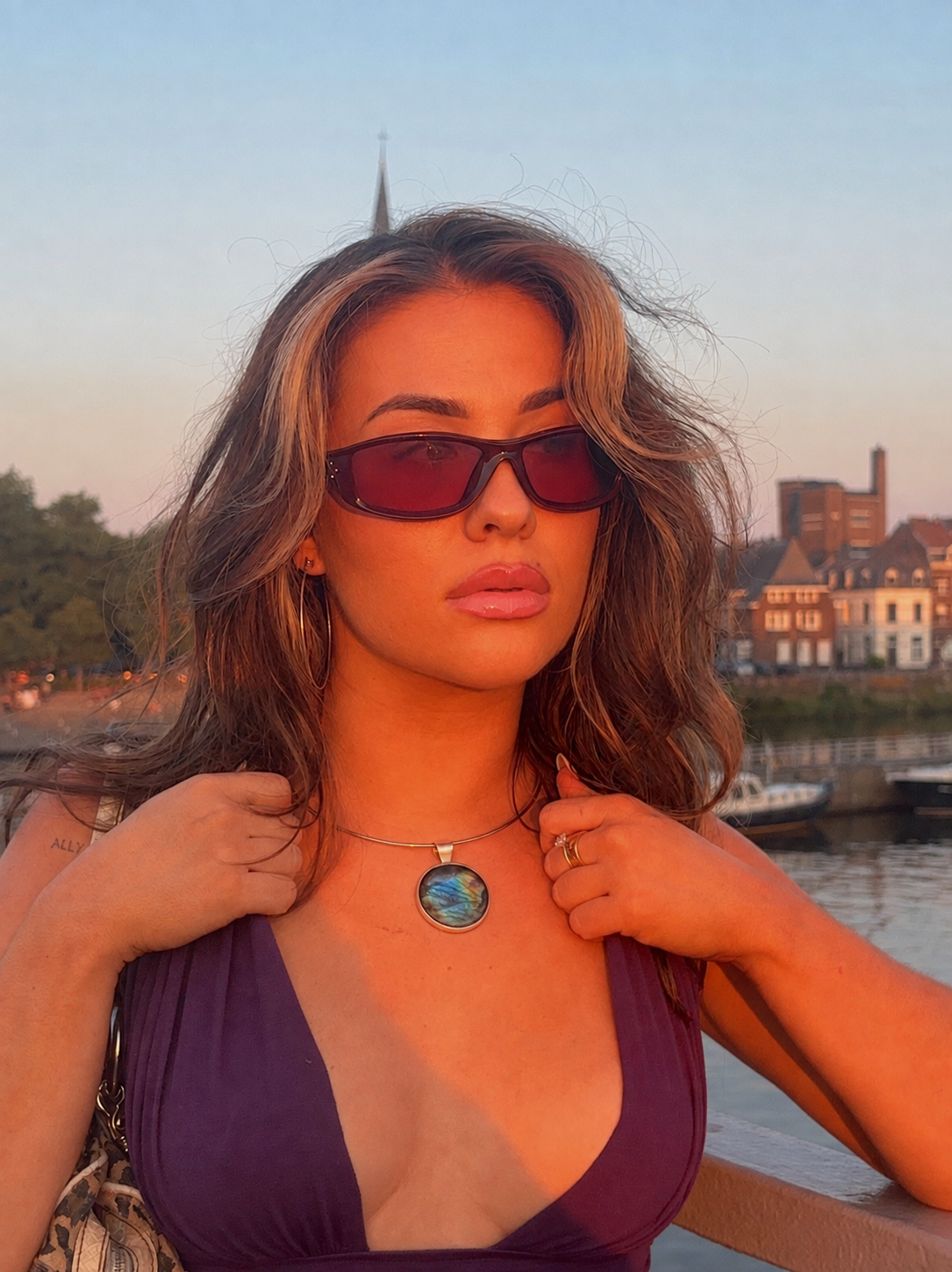 A woman outdoors by the water at sunset wearing dark tinted sunglasses, a purple bikini top, and a necklace with a round blue-green labradorite pendant.