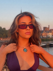 A woman outdoors by the water at sunset wearing dark tinted sunglasses, a purple bikini top, and a necklace with a round blue-green labradorite pendant.