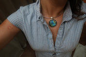 A close-up of a person wearing a silver-toned necklace with a round labradorite pendant featuring blue, green, and gold iridescent patterns, paired with a light blue and white striped button-down shirt.