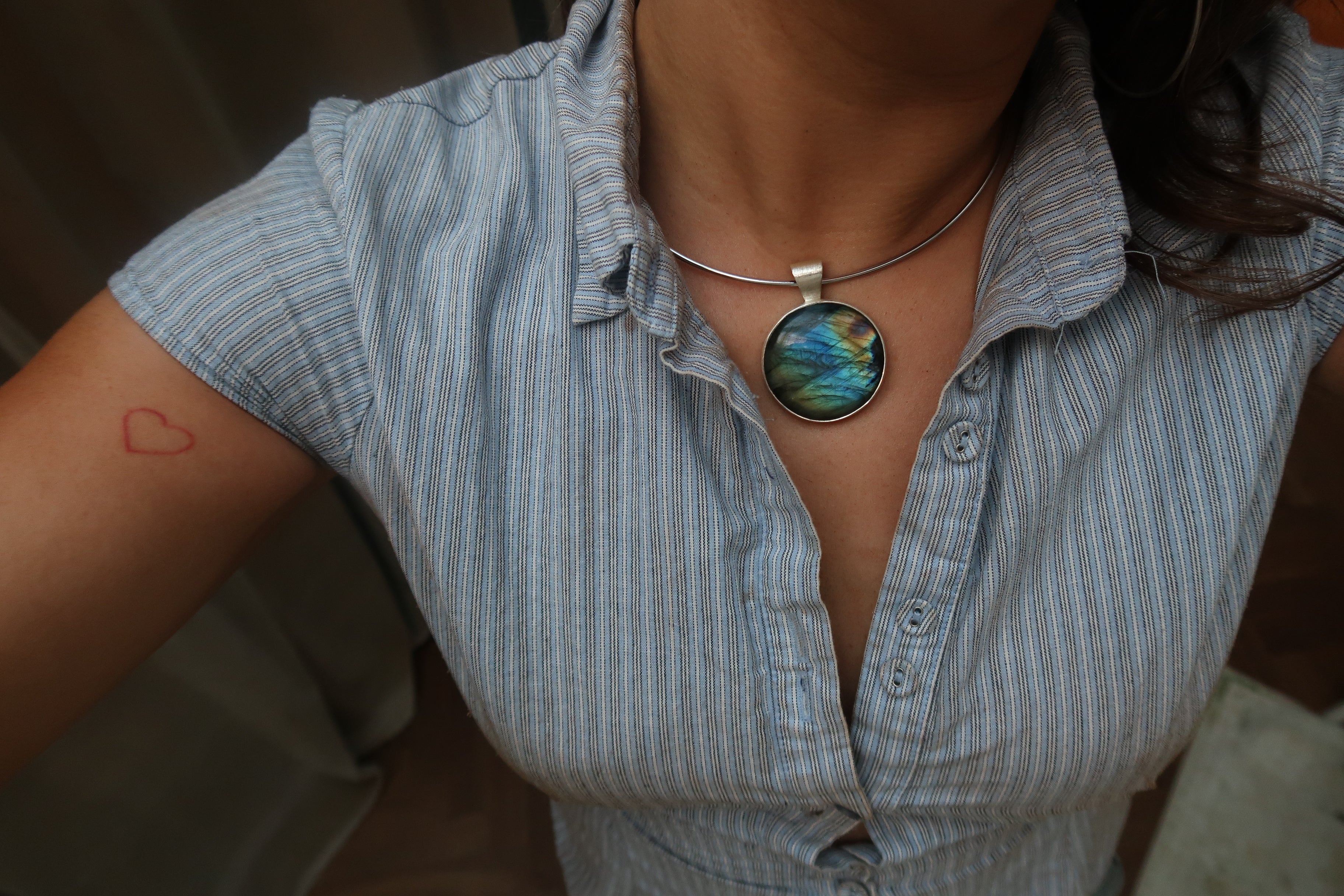 A close-up of a person wearing a silver-toned necklace with a round labradorite pendant featuring blue, green, and gold iridescent patterns, paired with a light blue and white striped button-down shirt.